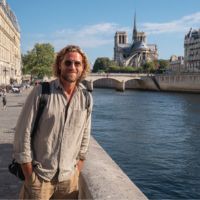 Bearded man in sunglasses and casual clothes leans on a stone railing by the Seine, Notre-Dame cathedral and bridge behind him.