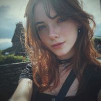 Young woman with auburn hair and freckles smiling softly in a selfie, rocky coastline and cloudy sky behind her.